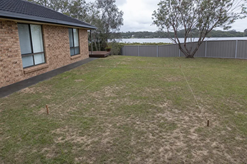 A string-line rectangle pegged out on a patchy backyard lawn behind a brick house where a pool is planned — pool excavation, Central Coast. Illustrative example.