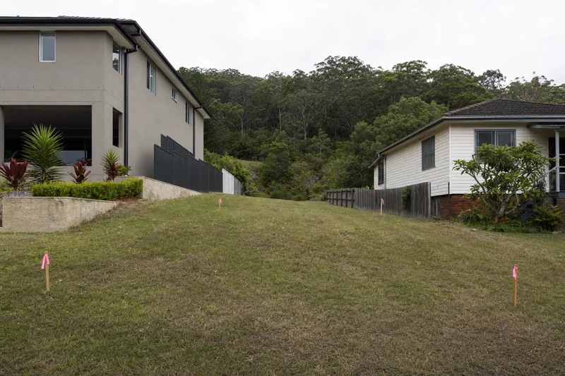 A vacant sloping block between two houses, mown grass with timber survey pegs tied with pink tape at its corners — site cuts, Central Coast. Illustrative example.