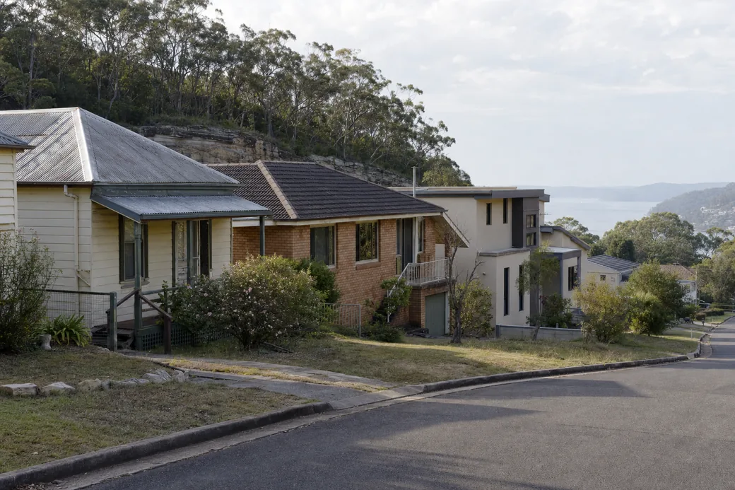 A hillside street of older and newer houses stepping down toward a wide stretch of water, sandstone and bushland above — excavation, Central Coast. Illustrative example.