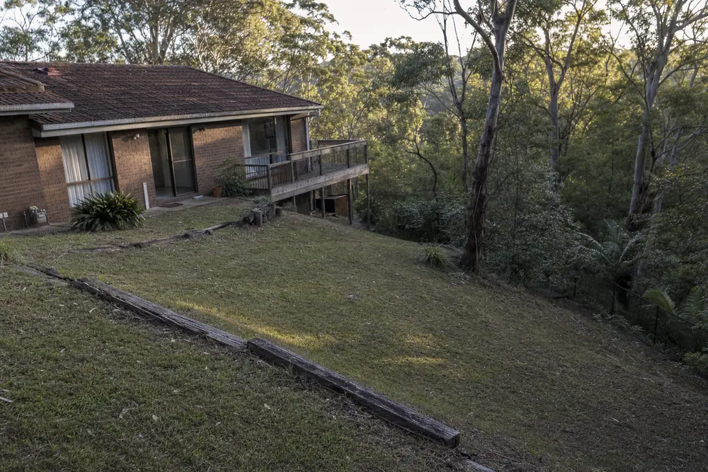 A steep grassed backyard falling away from the back of a brick-veneer house, old timber sleepers edging the slope, bushland behind in late sun — excavation, Central Coast. Illustrative example.