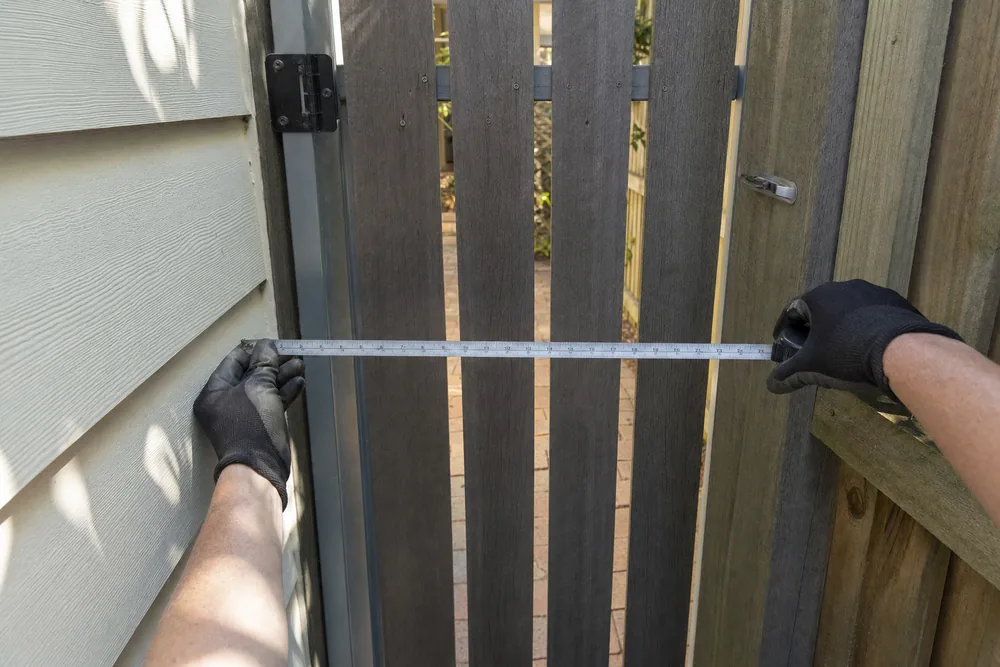 Gloved hands holding a tape measure across the width of a narrow timber side gate — tight access excavation, Central Coast. Illustrative example.